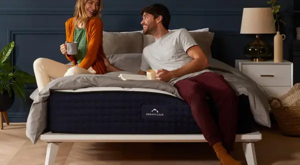 A couple enjoys coffee in bed. The DreamCloud hybrid mattress is on a white platform bed, answering the question: can you put a hybrid mattress on a box spring?