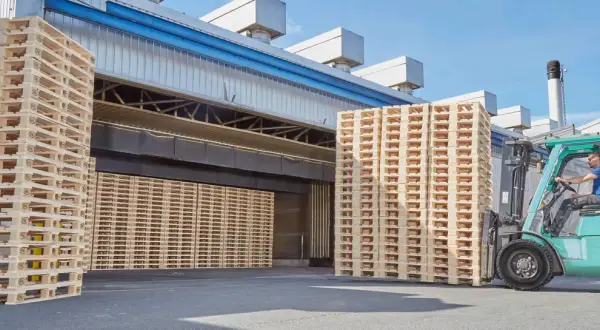 Un chariot élévateur déplace une pile de palettes traitées devant un entrepôt.