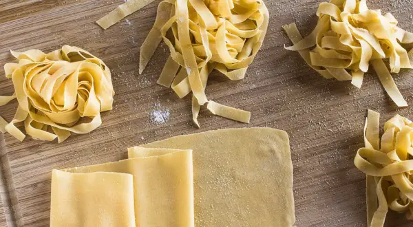 Frischer, selbstgemachter Pastateig, wie im original italienischen Rezept, liegt auf einem Holzbrett. Tagliatelle-Nester und Teigplatten warten auf ihre Zubereitung.