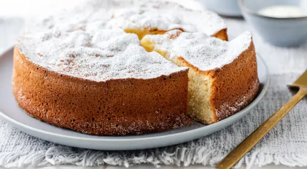 Una soffice torta allo yogurt, cosparsa di zucchero a velo, attende di essere gustata. Una fetta è già stata tagliata, rivelando il suo interno invitante.