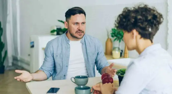 Man med intensiv blick förklarar något för kvinna, kanske om vad betyder narcissist. De sitter vid ett bord med kaffekoppar.