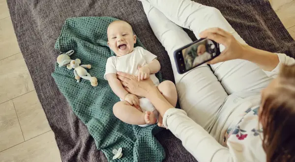 Uśmiechnięte niemowlę do kiedy będzie tak radosne? Mama robi mu zdjęcie telefonem, leży na kocyku z zabawkami.