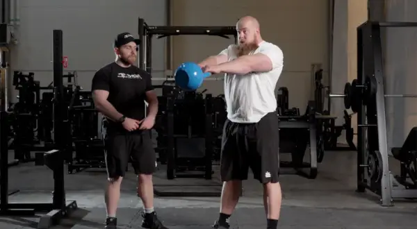 Två män i gym utför kettlebell swing. Den ena mannen, med skägg, håller i en blå kettlebell och är i rörelse. Den andra mannen står bredvid och observerar.