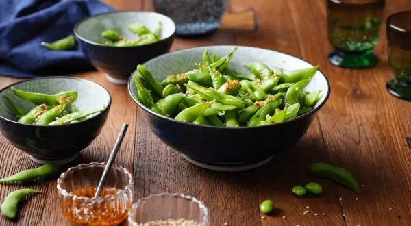 Schon gewusst, was ist Edamame? Grüne Sojabohnen in Schoten, gewürzt mit Sesam und Chili, serviert in Schalen auf Holztisch.