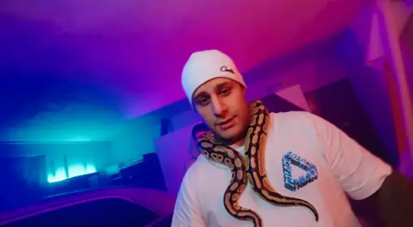 Macias w białej czapce i t-shircie z wężem na szyi, w neonowym świetle.