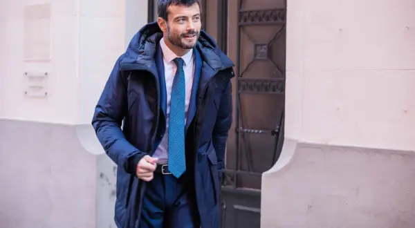 Un homme élégant porte un type de manteau homme bleu marine, un costume et une cravate, marchant dans la rue.