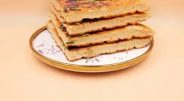 Stos kawałków omletu z odżywką białkową, udekorowany rozmarynem i fioletowym proszkiem, na talerzu.