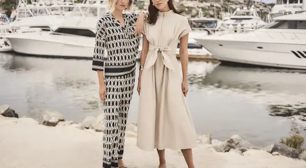 Dwie modelki prezentują kreacje z **joseph ribkoff katalog** na tle jachtów. Jedna w eleganckim, beżowym komplecie, druga w stylowym, wzorzystym stroju.