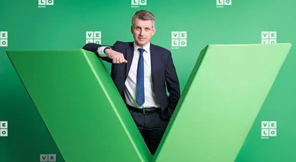 Mężczyzna w garniturze opiera się o zieloną literę "V" z logo Velo Bank.