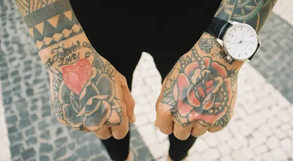 Dłonie pokryte kolorowym tatuażem tradycyjnym: róże, serce i napis "Forever Love". Na lewej ręce zegarek.