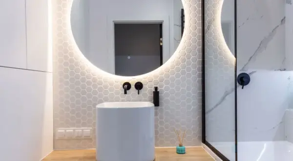 Moderno baño con lavabo blanco, espejo redondo iluminado y azulejos hexagonales. Ideal para tu presupuesto baño.