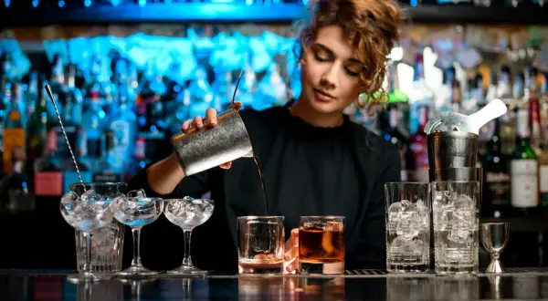 Barman nalewa drink z shakera do szklanki na tle podświetlonych butelek.