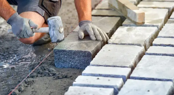 Układanie kostki brukowej na podsypce. Ręce w rękawiczkach kładą kamień, używając młotka. Widać suchy beton proporcje.