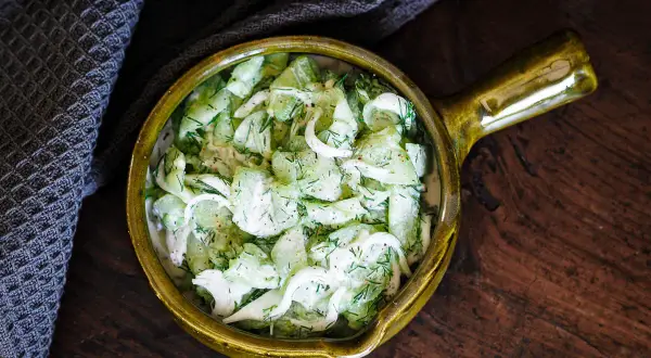 Salade de concombre à la crème, garnie d'aneth frais, servie dans un bol en céramique jaune sur une table en bois rustique.