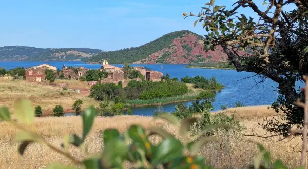 Vue sur le lac du Salagou, avec ses eaux bleues, ses collines rouges et les ruines d'un village. Un voilier navigue paisiblement.