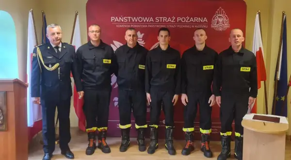 Sześciu strażaków w mundurach, w tym dowódca, tworzy rota strażacka. Stoją na tle sztandarów i logo Państwowej Straży Pożarnej.