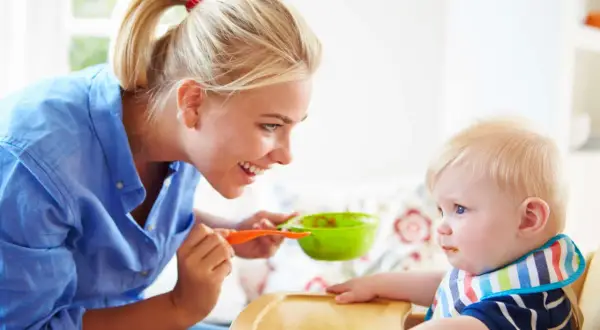 Mama karmi dziecko łyżeczką z zielonej miseczki. Dziecko ma śliniaczek, a mama uśmiecha się do niego. To posiłek bogaty w błonnik dla dziecka.