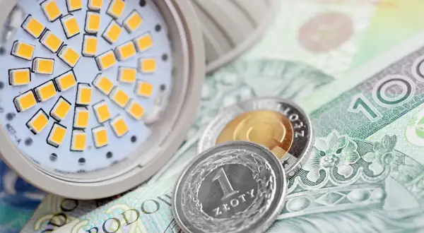 Żarówka LED i monety na banknocie 100 zł. Symbolizuje to wniosek o zamrożenie cen prądu dla gospodarstw domowych.
