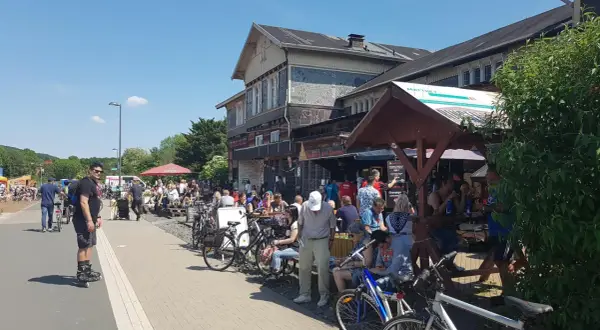 Menschen genießen das Leben am **Wuppertaler Hauptbahnhof**. Viele sitzen draußen, einige mit Fahrrädern, bei Sonnenschein.