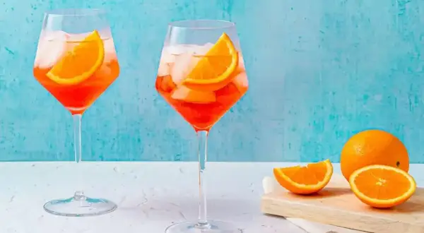 Två glas med Aperol Spritz, is och apelsinskivor, med en hel apelsin och skivor på en skärbräda i bakgrunden.