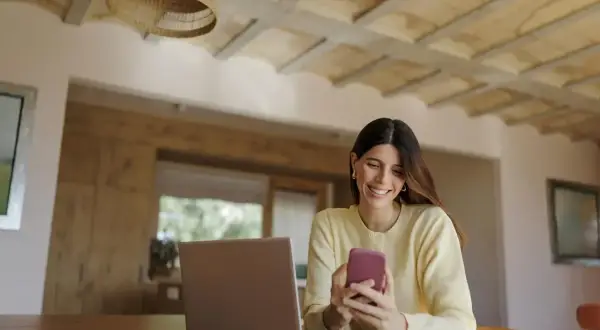 Uśmiechnięta kobieta korzysta z telefonu, planując przelewy w mBanku. Obok laptop i notatnik.