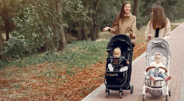 Dwie kobiety spacerują z dziećmi w wózkach po parku jesienią.