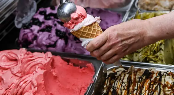 Eine Hand füllt eine Waffel mit rosa Eis. Daneben stehen Behälter mit verschiedenen Eissorten, darunter Blaubeer- und Pistazieneis. Das beste Eis der Welt!