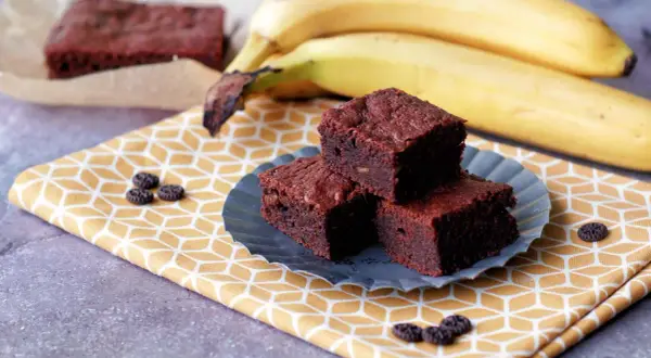 Saftige Bananen-Brownies auf einem gelben Tuch, daneben reife Bananen. Ein Genuss!
