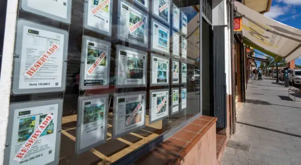 Escaparate de inmobiliaria con anuncios de venta de casas, muchos marcados como "RESERVADO". Se vislumbra una casa señorial en uno de los carteles.