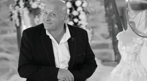 Jacek, znany jako królowe życia, w eleganckim garniturze, z uśmiechem spogląda w obiektyw. W tle widać suknię ślubną.