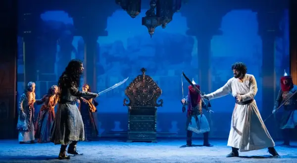 Escena del musical "El Médico" en Zaragoza. Dos hombres se enfrentan con espadas ante un trono dorado, rodeados por otras figuras y un fondo azul.