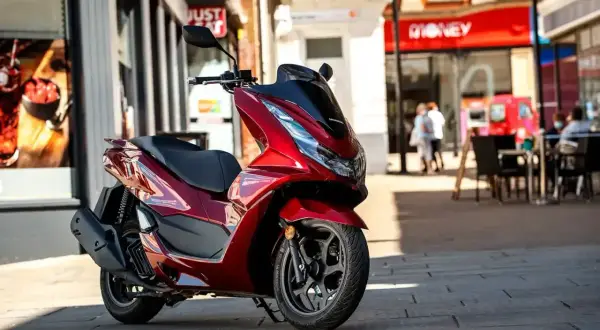 Czerwony skuter Honda PCX 125 zaparkowany na chodniku przed sklepami.
