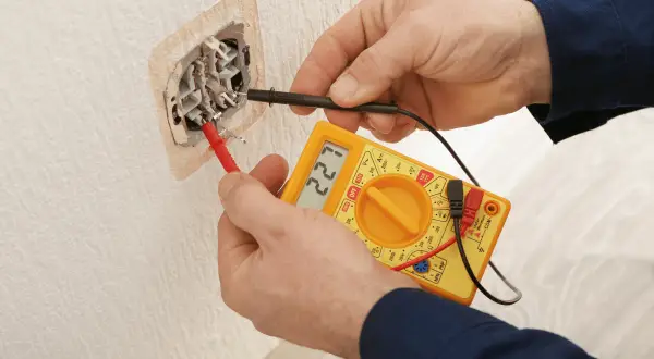 Elektryk sprawdza gniazdko miernikiem. Na wyświetlaczu widnieje 221V, co oznacza, że napięcie jest w normie.