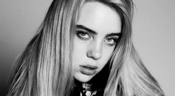 Zbliżenie na twarz Billie Eilish, artystki znanej z unikalnych utworów. Jej spojrzenie jest intensywne, a długie włosy opadają na ramiona.