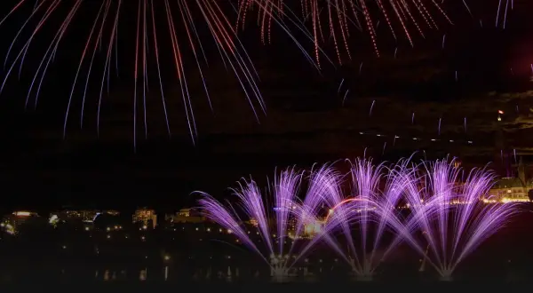 Feu d'artifice violet et blanc illuminant un ciel nocturne au-dessus d'une ville et de bâtiments historiques.