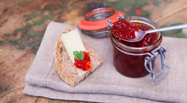 Un cucchiaino raccoglie la lucida marmellata di peperoni da un barattolo. Sul pane, formaggio e un tocco di questa delizia.