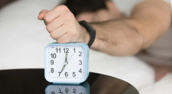 A hand rests on an alarm clock showing 7:00, reflecting the question: is four hours of sleep enough?