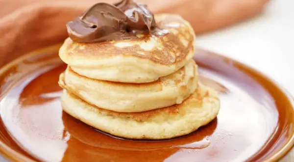 En hög med fluffiga amerikanska pannkakor toppas med chokladkräm på en brun tallrik.