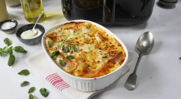 En gyllene lasagne i airfryer, toppad med smält ost och färsk basilika. Serveras med en sked och tillbehör som olivolja och parmesan.