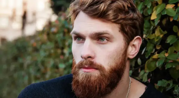 Un homme au visage ovale, avec une barbe rousse et des yeux bleus perçants, regarde au loin, entouré de verdure.