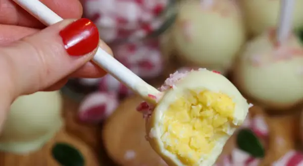 En hand med rött nagellack håller en cake pop med vit choklad och krossade polkagrisar. Perfekt för ett cake pops recept!