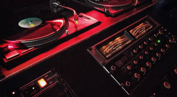 Giradischi e mixer in un locale notturno, illuminati da luci rosse. Il logo "BASIC CLUB" è visibile su uno dei giradischi.