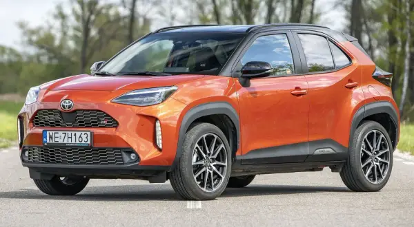 Pomarańczowy Toyota Yaris Cross 2024 z czarnym dachem, zaparkowany na drodze, z drzewami w tle.