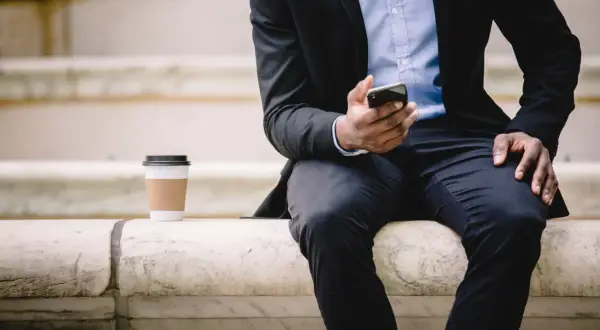 Mężczyzna w garniturze siedzi na kamiennych schodach, trzymając telefon. Kawa obok. Czeka na ważne powiadomienie, może to być przekierowanie na nowe zadanie.