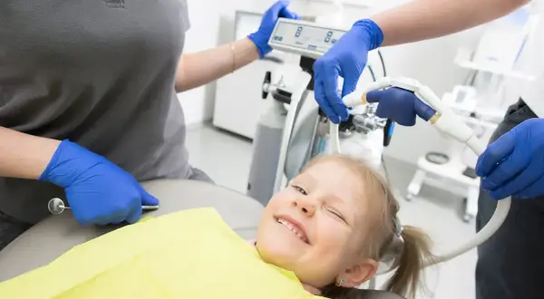Uśmiechnięta dziewczynka na fotelu dentystycznym, stomatolog przygotowuje sprzęt.