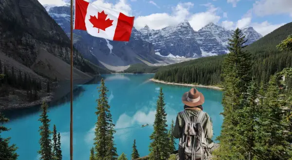 Persona con mochila admira el lago turquesa y las montañas nevadas de Canadá. La bandera canadiense ondea, invitando a una aventura de work and holiday Canada.