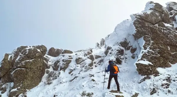 Un excursionista asciende una ladera nevada. Las rocas cubiertas de nieve forman una silueta que recuerda a una montaña con forma de mujer.