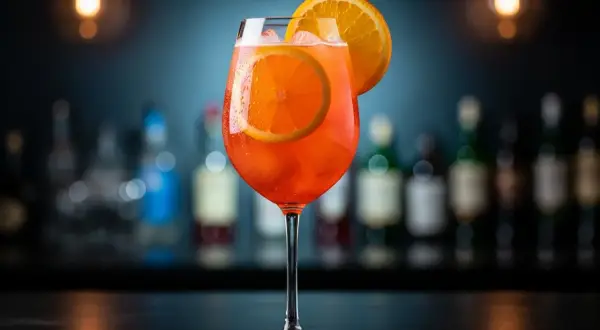 Ett glas med en orange aperol drink utan prosecco, garnerad med apelsinskivor och is. Bakom syns en barmiljö med flaskor.