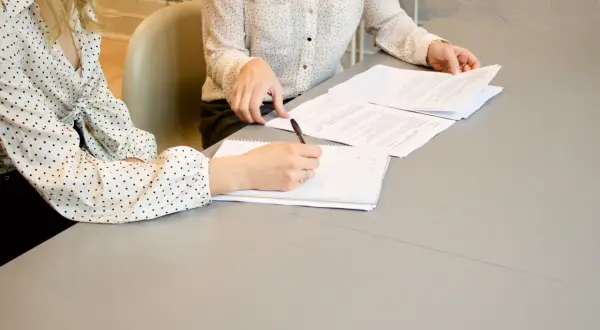 Dwie kobiety przy stole analizują dokumenty. Jedna pisze notatki, druga przegląda papiery. Komparycja ich pracy jest widoczna.