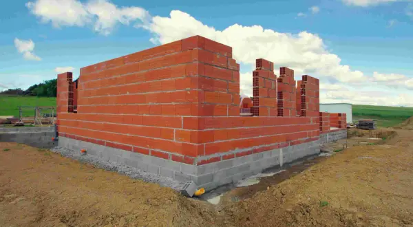 Budowa domu 100m2 w toku. Czerwone ściany z pustaków, fundamenty z bloczków betonowych, niebo z chmurami.